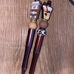 May include: Two brown and black pens with decorative tops. One top is a brown coffee cup with the word "Coffee" on it. The other top is a brown and yellow iced coffee cup with a pink straw.