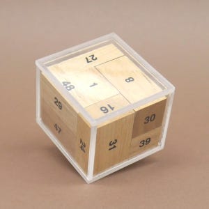 Può includere: Un cubo puzzle in legno con una custodia in plastica trasparente. Il cubo è composto da blocchi di legno con numeri neri. Il legno è marrone chiaro. Il cubo è appoggiato su una superficie marrone.