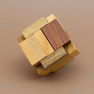 Könnte beinhalten: Ein hölzerner Block-Puzzle-Würfel, der aus mehreren rechteckigen Teilen in verschiedenen Holztönen besteht, darunter helles Beige, mittleres Braun und ein dunkleres Rotbraun. Die Teile sind ineinandergreifend zu einer festen, dreidimensionalen Form.