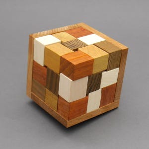 Può includere: Un puzzle a cubo in legno con varie tonalità di legno, tra cui marroni chiari e scuri, arancione e bianco. Il puzzle è composto da piccoli blocchi quadrati disposti a formare un cubo più grande.