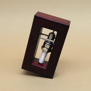 May include: A spark plug is displayed in a dark red wooden frame. The frame is tilted to the side, and the spark plug is visible through the opening in the frame.