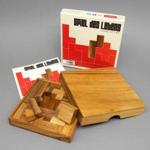 Könnte beinhalten: Ein Holzbox-Puzzle namens "Spiel des Lebens" oder "Game of Life". Die Box ist geöffnet und die Holzpuzzle-Teile liegen lose darin. Der Boxdeckel ist aus Holz und hat einen kleinen Ausschnitt an der Vorderseite. Die Box ist mit "Spiel des Lebens" und "Game of Life" in schwarzer Schrift auf weißem Hintergrund beschriftet.