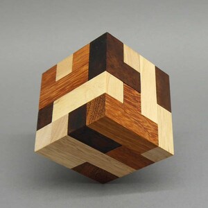 Könnte beinhalten: Ein Holzkubus-Puzzle mit einem komplexen Design. Der Würfel besteht aus verschiedenen Holzarten in den Farben Hellbraun, Dunkelbraun und Schwarz. Das Puzzle ist so zusammengesetzt, dass ein geometrisches Muster entsteht.