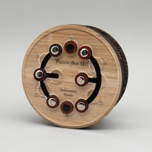 May include: A wooden Puzzle-Box 11 with a clear acrylic face. The puzzle has a circular design with black accents, wooden and silver-colored knobs. The text "Siebenstein Spiele" is also visible.