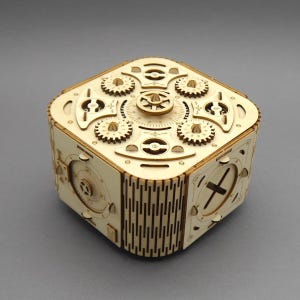 Op de afbeelding: Een lichtgekleurde houten puzzeldoos met een complex mechanisch ontwerp. Het bovenoppervlak toont meerdere in elkaar grijpende tandwielen, ronde wijzerplaten en ingewikkelde uitsparingen. De zijpanelen vertonen geometrische patronen en draaimechanismen.