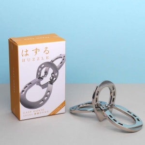 Könnte beinhalten: Ein silbernes Metallpuzzle in Form eines Hufeisens. Das Puzzle befindet sich in einer Schachtel mit gelbem Hintergrund und japanischem Text, der "hazur" und "puzzle" lautet. Die Schachtel trägt außerdem die Aufschrift "Cast Horse" und "Level *****".