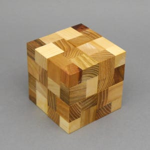 Op de afbeelding: Een houten puzzelkubus bestaande uit kleinere in elkaar grijpende blokjes. De kubus heeft een verscheidenheid aan natuurlijke houttinten, variërend van licht crème tot donkerbruin, met duidelijke houtnerfpatronen op elk vierkant oppervlak.