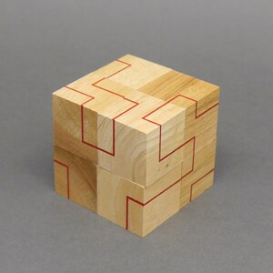Puede incluir: Un rompecabezas de cubo de madera con líneas rojas que forman un patrón geométrico en cada lado. El cubo está hecho de ocho cubos más pequeños que se pueden reorganizar para crear diferentes patrones.