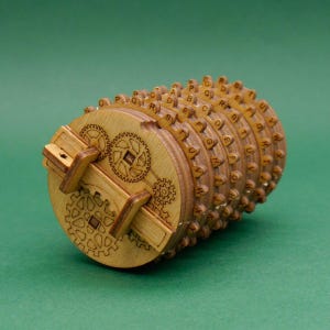 Könnte beinhalten: Ein zylindrisches Kryptex-Puzzle aus Holz mit mehreren rotierenden Ringen, die jeweils mit Buchstaben des Alphabets graviert sind. Das kreisförmige Endstück zeigt komplizierte Zahnradmuster und einen Schließmechanismus aus hellem Holz.
