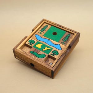Könnte beinhalten: Eine Holzkästchen-Puzzle mit einem Golfplatz-Design. Das Puzzle zeigt einen grünen, blauen und gelben Golfplatz mit Bäumen und einem Loch. Die Box ist aus dunklem Holz und hat einen Klappdeckel.
