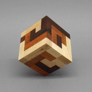 Può includere: Un puzzle di cubo di legno con un motivo geometrico realizzato con pezzi di legno di diversi colori. Il cubo è fatto di legno chiaro, legno scuro e legno marrone.