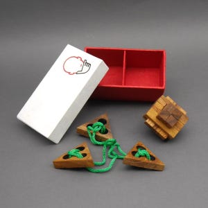 Könnte beinhalten: Eine weiße Box mit rotem Innenraum und roter Grafik, neben Holzpuzzles. Die Box hat eine rote Grafik eines Kopfes und einer Hand. Drei Holz-Dreiecke sind durch eine grüne Schnur verbunden. Ein Holz-Würfel-Puzzle ist ebenfalls vorhanden.