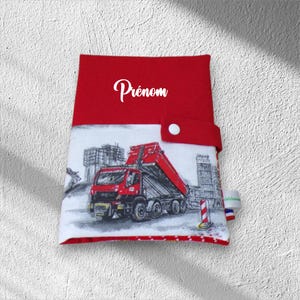 Peut inclure: Une pochette en tissu rouge avec une fermeture à pression blanche et le mot Prénom imprimé en blanc. La partie inférieure présente une illustration graphique d'un camion benne rouge sur un chantier avec des bâtiments gris en arrière-plan.