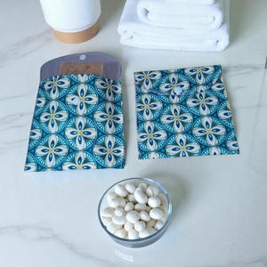 Peut inclure: Deux pochettes à motifs turquoise et blancs avec fermeture à bouton-pression sont présentées à côté d'un petit bol en verre rempli de bonbons blancs. Une pile de serviettes blanches et une plante en pot sont également dans l'image.