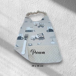 Peut inclure: Un bavoir beige avec un motif de véhicules de chantier, une section inférieure à pois blancs et le texte Prénom. Le bavoir possède une encolure élastique et mesure 29 x 36 cm. Une fourchette argentée à manche blanc est posée sur le bavoir.