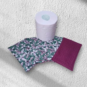 Peut inclure: Un rouleau de papier toilette blanc placé derrière plusieurs lingettes rectangulaires en tissu. Les lingettes présentent un fond vert avec un motif répétitif de papillons blancs et roses, tandis qu'une lingette est magenta foncé uni texturé.