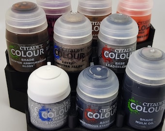 May include: A set of ten Citadel Colour acrylic paints in various shades of gray, brown, and black. The paints are in small bottles with clear lids and are arranged in a circular holder. The labels on the bottles include the paint name, shade, and brand.