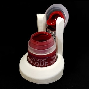 Puede incluir: Soporte para botes de pintura blanco impreso en 3D con un bote de pintura Citadel Colour rojo. El soporte tiene una tapa que se levanta para permitir el acceso al bote de pintura.