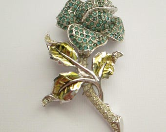 May include: A silver tone brooch in the shape of a rose with green and gold leaves. The rose is covered in clear and light blue rhinestones.