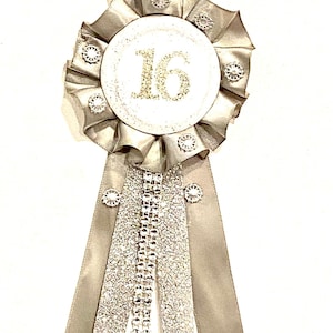 May include: A silver ribbon rosette badge featuring a central white circular disc with the number 16 in gold glitter. The pleated silver satin ribbon is accented with small circular rhinestone embellishments and two long silver glitter fabric streamers.