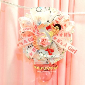 May include: A decorative baby shower favor featuring a pink and white ribbon bow with the text "It's A Girl". The bow is adorned with a baby-themed embellishment and a pink rose. The favor is attached to a clear glass container.