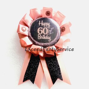 May include: A circular rosette badge featuring a black center button with the text Happy 60th Birthday in rose gold glitter. The badge is surrounded by pleated light pink ribbon, accented with small rose gold studs, and two black glittery ribbon tails.