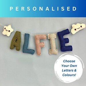 May include: A crocheted name banner with the name "ALFIE" spelled out in different colored letters. The letters are strung together with a white string and there are two small crocheted characters on either end of the banner. The text "Choose Your Own Letters & Colours!" is written in a white circle on a blue background.