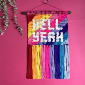 Op de afbeelding: Een regenboogkleurig gehaakt wandkleed met de woorden "HELL YEAH" in wit garen. Het kleed heeft een houten lat aan de bovenkant en kleurrijke franjes aan de onderkant.