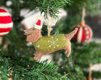 Puede incluir: Un adorno de dachshund de fieltro con un suéter verde con un diseño de copo de nieve y un gorro de Papá Noel rojo colgado de un árbol de Navidad verde.