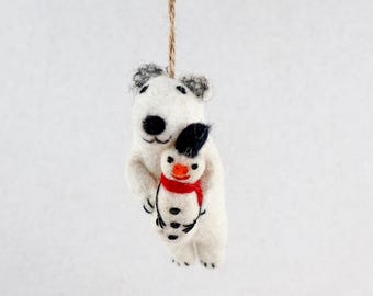 May include: A white felted polar bear ornament hanging from a brown twine string. The polar bear has black eyes and a black nose, and it is holding a small snowman figure that features a black hat, an orange nose, and a red scarf around its neck.