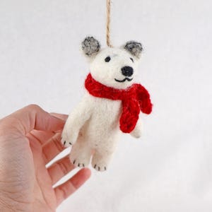 以下が含まれることがあります： 白い羊毛フェルトで作られた小さなホッキョクグマのオーナメント。鮮やかな赤のかぎ針編みのマフラーを巻き、黒い刺繍の目と鼻、グレーの耳先が特徴です。頭頂部には細いライトブラウンの麻紐が付いています。
