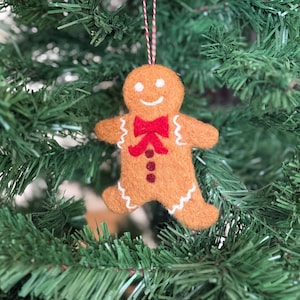 Könnte beinhalten: Ein Filz-Lebkuchenmann-Weihnachtsschmuck mit roter Fliege und drei roten Knöpfen. Der Schmuck hat weiße Nähte und ein lächelndes Gesicht. Er hängt an einer rot-weißen Schnur an einem grünen Weihnachtsbaum.