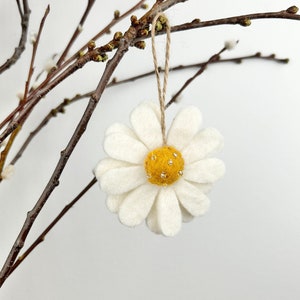 Könnte beinhalten: Ein weißes Filz-Gänseblümchen-Ornament mit gelber Mitte und goldenen Perlenakzenten, das an einer Schnur aus Naturfaser hängt. Das Gänseblümchen hängt vor braunen Ästen mit Knospen, was ein Frühlingsmotiv suggeriert.