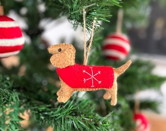 Puede incluir: Un adorno de dachshund de fieltro que lleva un suéter rojo con un diseño de copo de nieve blanco. El adorno está colgado de un árbol de Navidad verde.