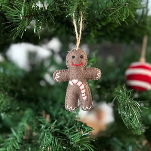 Könnte beinhalten: Ein brauner Filz-Lebkuchenmann-Anhänger mit einem weißen Zuckerstangen-Design, der an einem grünen Weihnachtsbaum hängt.