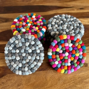 Op de afbeelding: Vier ronde vilt onderzetters, twee zijn grijs en twee zijn veelkleurig met een verscheidenheid aan felle kleuren. De onderzetters zijn gemaakt van kleine viltballen.