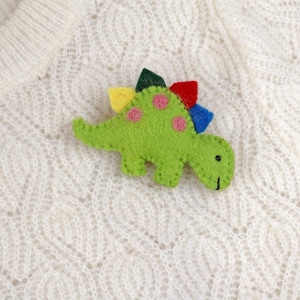 Pode incluir: Um broche de dinossauro em feltro na cor verde-lima brilhante com costuras verde-escuras visíveis nas bordas. O dinossauro apresenta manchas circulares cor-de-rosa nas costas e uma fileira de espinhos triangulares em amarelo, vermelho e azul.