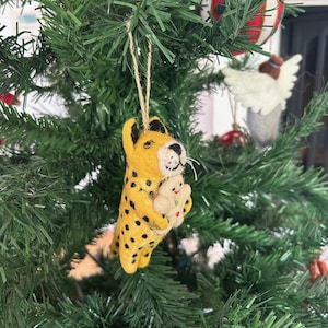 Könnte beinhalten: Ein gelber und schwarzer Filz-Geparden-Anhänger, der ein Lebkuchen-Keks hält. Der Anhänger hängt an einem grünen Weihnachtsbaum.