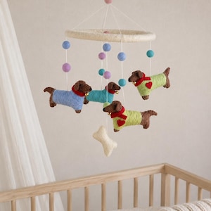 May include: A hanging mobile featuring four felted brown dachshunds wearing sweaters in light blue, teal, and lime green with red heart accents and small gold bells. The mobile includes colourful felt balls and a cream-coloured felt bone shape.