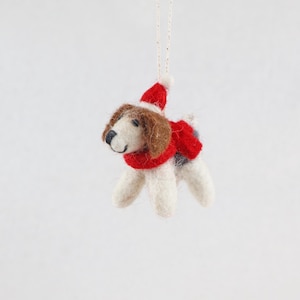 Könnte beinhalten: Ein aus Filzwolle gefertigter Hundeanhänger mit braunem und weißem Fell, der einen roten Schal und eine passende rote Weihnachtsmütze mit weißem Bommel trägt. Die kleine Hunde-Figur hängt an einer dünnen, hellen Schnur an der Mützenspitze.