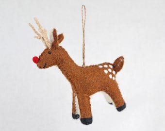 May include: A brown felt reindeer ornament featuring a bright red nose, light tan antlers, and white spots on its back. The reindeer has black hooves and a jute twine loop attached to the top for hanging, with visible stitching along the edges.