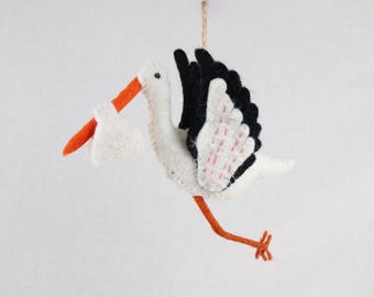 May include: A felt stork ornament featuring a white body, black wing feathers, and orange beak and legs. The stork carries a small white bundle in its beak and is suspended by a thin brown string attached to the top of its back for hanging.