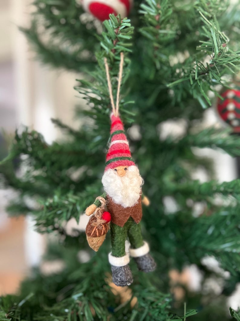 May include: A felt gnome ornament with a red and white striped hat, brown beard, and green pants. The gnome is holding a brown acorn with a red cap.