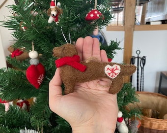 Puede incluir: Un adorno de dachshund de fieltro marrón con una bufanda roja y un corazón blanco con un diseño de copo de nieve rojo. El adorno está colgado de un árbol de Navidad.