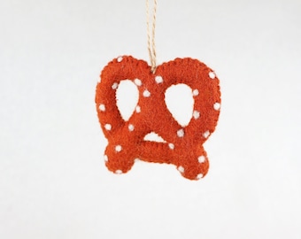 May include: A felt pretzel ornament in a burnt orange color with small white embroidered dots scattered across the surface. The pretzel is constructed with visible stitching along the edges and includes a thin tan twine loop for hanging purposes.