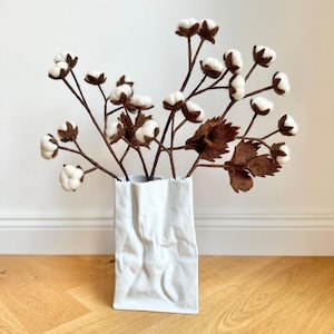Könnte beinhalten: Eine weiße Keramikvase in Form einer Papiertüte mit einem Strauß weißer Baumwollblütenstängel mit braunen Zweigen.
