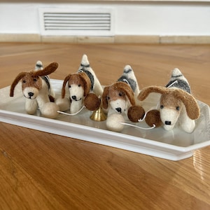 Könnte beinhalten: Vier Filz-Hundefiguren auf einem weißen Tablett. Die Hunde sind braun und weiß mit schwarzen Markierungen. Sie sind durch eine Schnur mit einer kleinen goldenen Glocke in der Mitte verbunden.