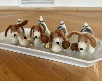 May include: Four felt dog figurines on a white tray. The dogs are brown and white with black markings. They are connected by a string with a small gold bell in the middle.