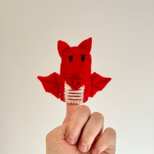 May include: Red felt finger puppet shaped like a dragon with white and red stripes on the neck. The dragon has black eyes and a grumpy expression.