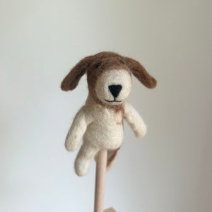 May include: A handmade felt dog figurine with brown and white coloring. The dog has floppy ears, a black nose, and is mounted on a wooden stick. The figurine is a whimsical decorative item.